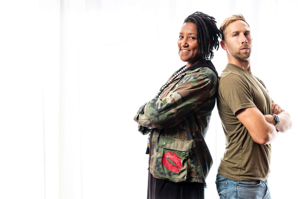 Miki Vale and Nick Isabella standing back to back with arms crossed, on a white background. Miki on the left wearing a camo jacket and Nick on the right wearing a brownish green shirt. They present as a power duo for this month's About The Cover.