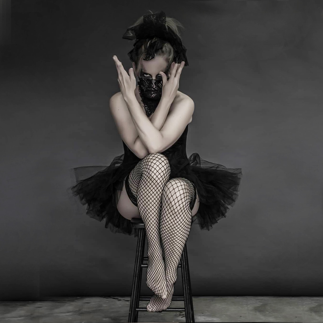 Ballerina sitting on a high chair, wearing a black tutu skirt with black corsage; covering her mouth with a screened scarf and with both arms crossed near her face forming a V