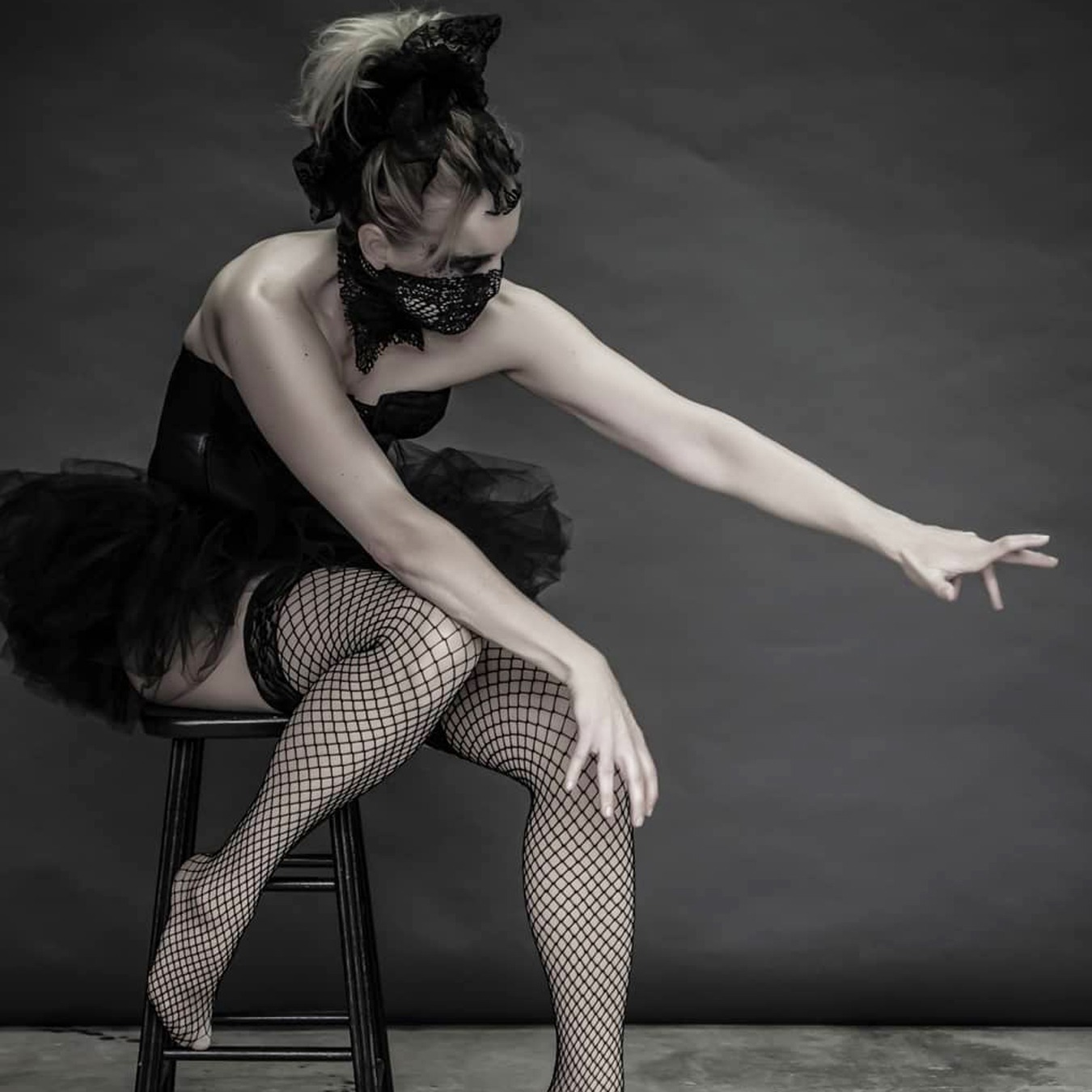 Ballerina sitting on a high chair with her left foot on the ground, wearing a black tutu skirt with black corsage; covering her mouth with a screened scarf and looking likes she is trying to reach something on the ground