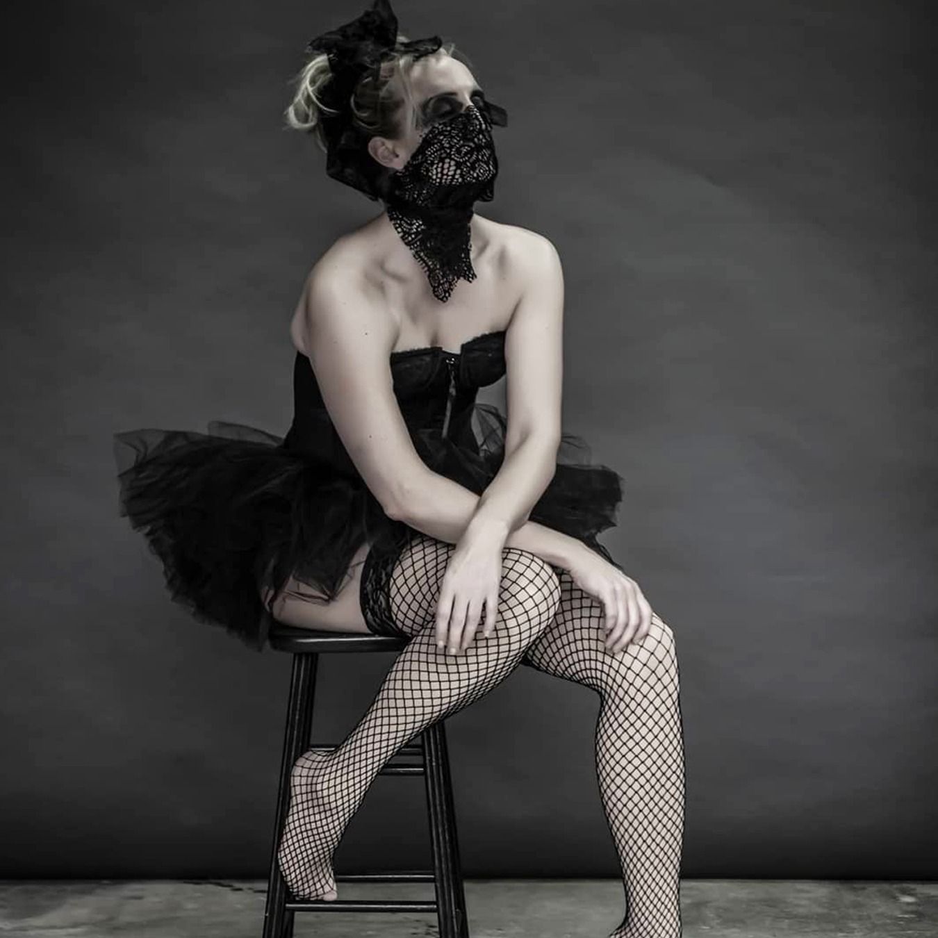 Ballerina sitting on a high chair with her left foot on the ground, wearing a black tutu skirt with black corsage; covering her mouth with a screened scarf looking up like waiting for something to happen