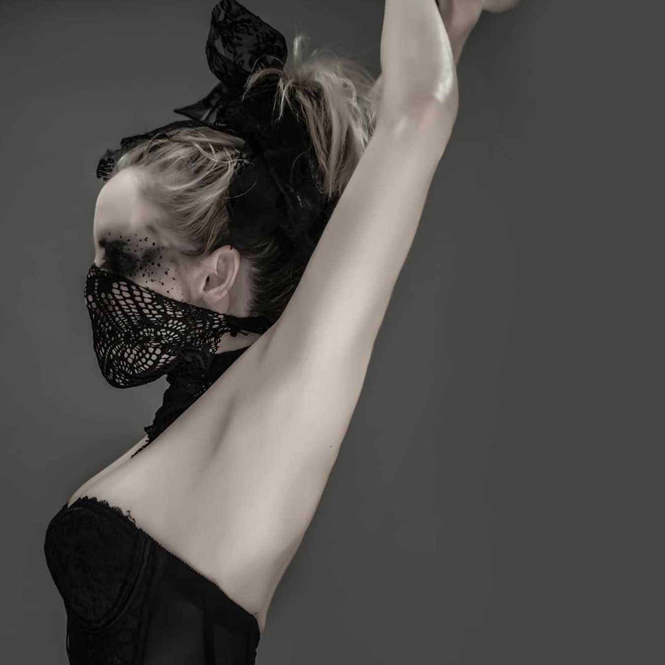 Ballerina wearing a black tutu skirt, black corsage and with her mouth partially covered by a screened scarf stand on a ballet position looking left with both her arms on the air