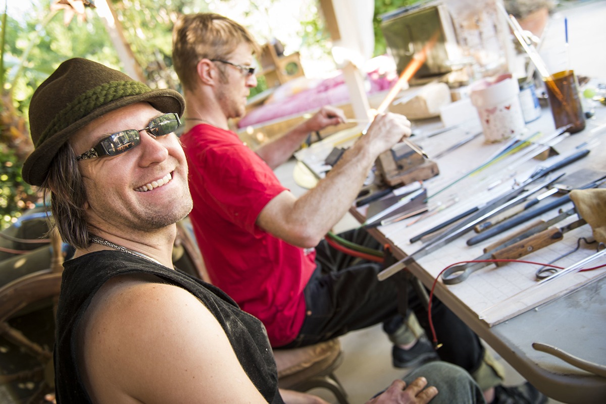 Glass artists Corey Mitchell smiling at the camera while Alexander Chacona is working very concentrated in the background; glass tube on the flame taking it's next form