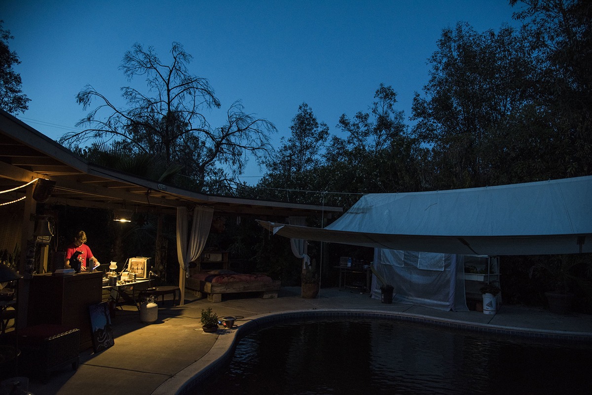 Beautiful open shot framing a blue sky while glass artist Alexander Chacona is working under a lamp, by the pool on his work station for the day.