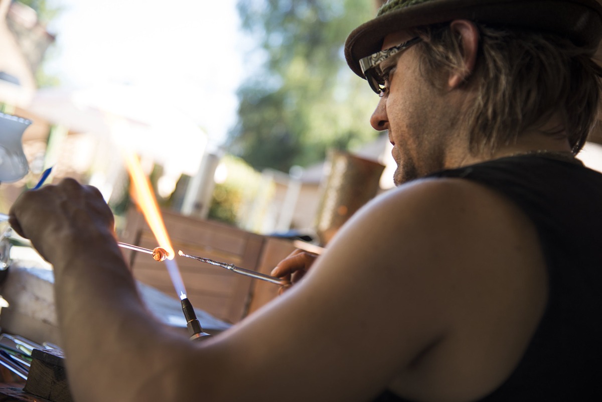 Glass artist Corey Mitchell sitting on the right of the frame, holding two pieces of glass, almost touching themselves right at center on the flame of the blow torch.