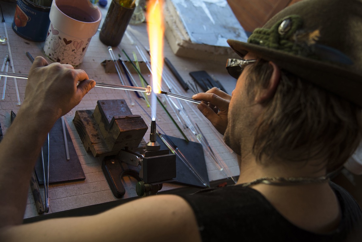 Aerial shot of glass artist, Corey Mitchell doing his art; tool set on the table, a couple of thin glass tubes and a furious flame forming and molding the piece of glass.
